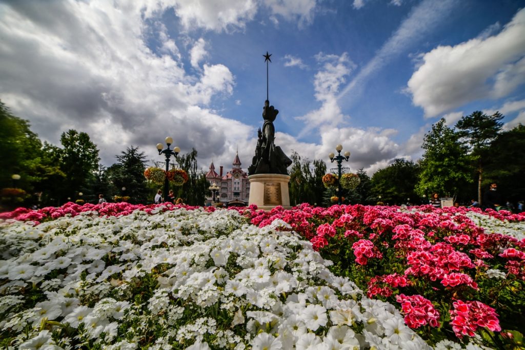 Un parterre de fleur, au centre une sculpture représentant un parchemin et la main de Mickey tenant une baguette magique