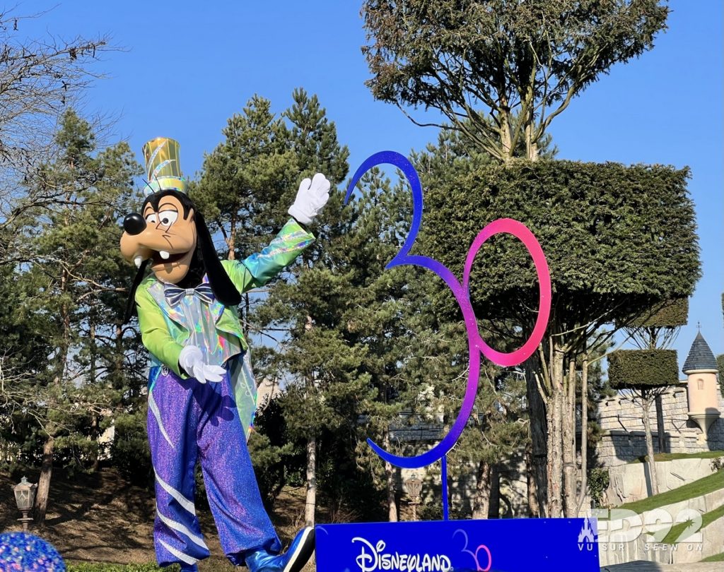 Dingo pose à côté du logo des 30 ans de Disneyland Paris. Il est sur la colline devant le château de la belle au bois dormant. 